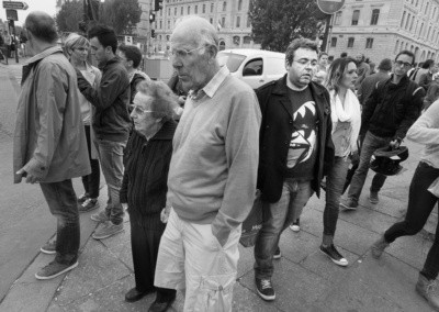Photo shows an elderly couple waiting to cross a street corner as a young man passes behind them wearing a scary looking t-shirt.
