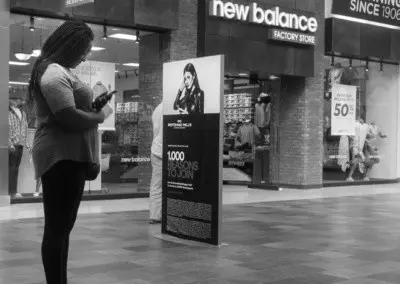 Photo shows a woman looking at her phone while standing in front of a billboard that has a woman on it looking at her phone.