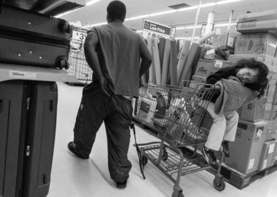 Photo shows a young girl resting her head on a blanket that covers the hand bar of a shopping cart as if she was falling asleep. Dad pulls the cart as the heavy weight of his key chain pulls his pants down.