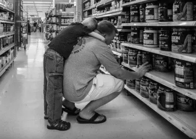 Photo shows a young boy with his arms wrapped around his father who is looking for protein mixes in a department store.