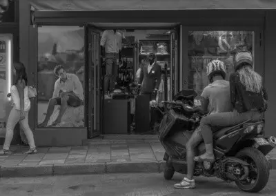 Photo shows a woman walking down the sidewalk as a shoppe mannequin appears to be doing a second take as it leans to see around the doorway. At the same time, a photo of a male model appears to be looking at a guy or girl on a motor scooter that just arrived.