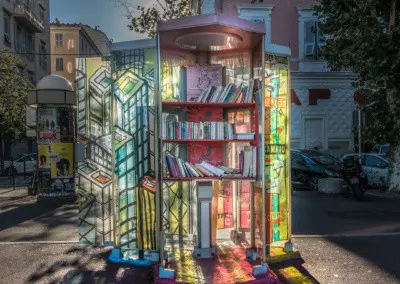 Photo shows an old french telephone booth that has been converted to a book borrow station. Sunlight comes through the hand colored glass that reflects onto the ground.